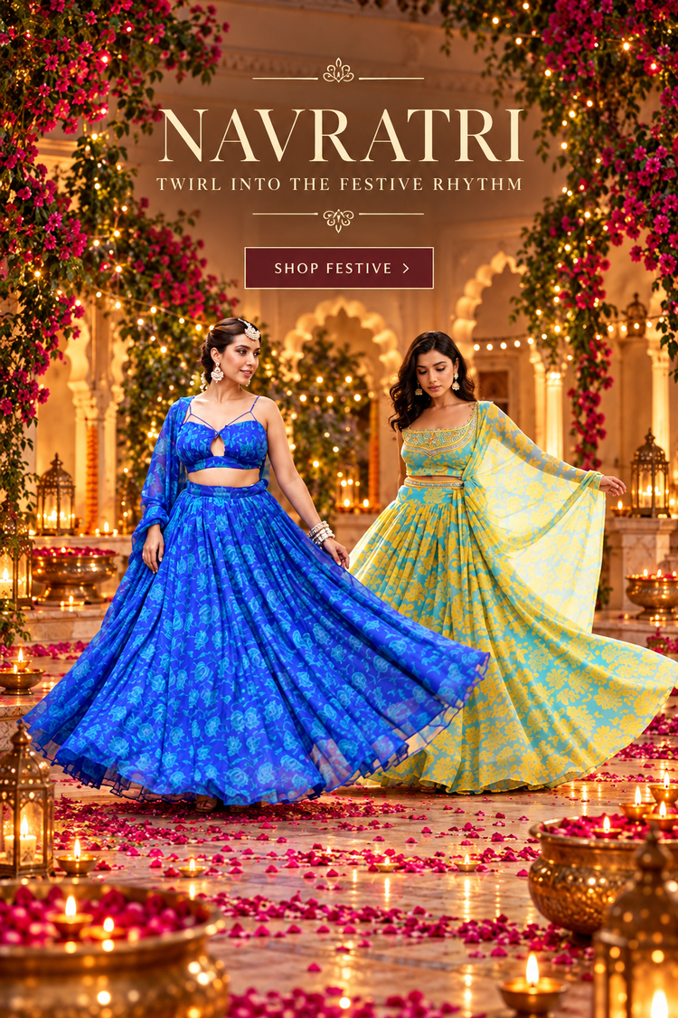 Two women modeling traditional Indian lehenga gharara suits in a festive indoor setting decorated with hanging pink flowers, diyas, and rose petals. One model wears a royal blue patterned suit, and the other wears a yellow and green patterned suit.